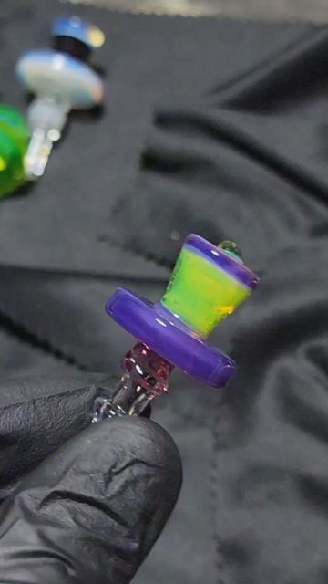 Gem Cup Gem-Lock cap held in a black glove: a purple and lime-green hat-shaped glass cap with a purple base ring, more colored caps blurred behind it